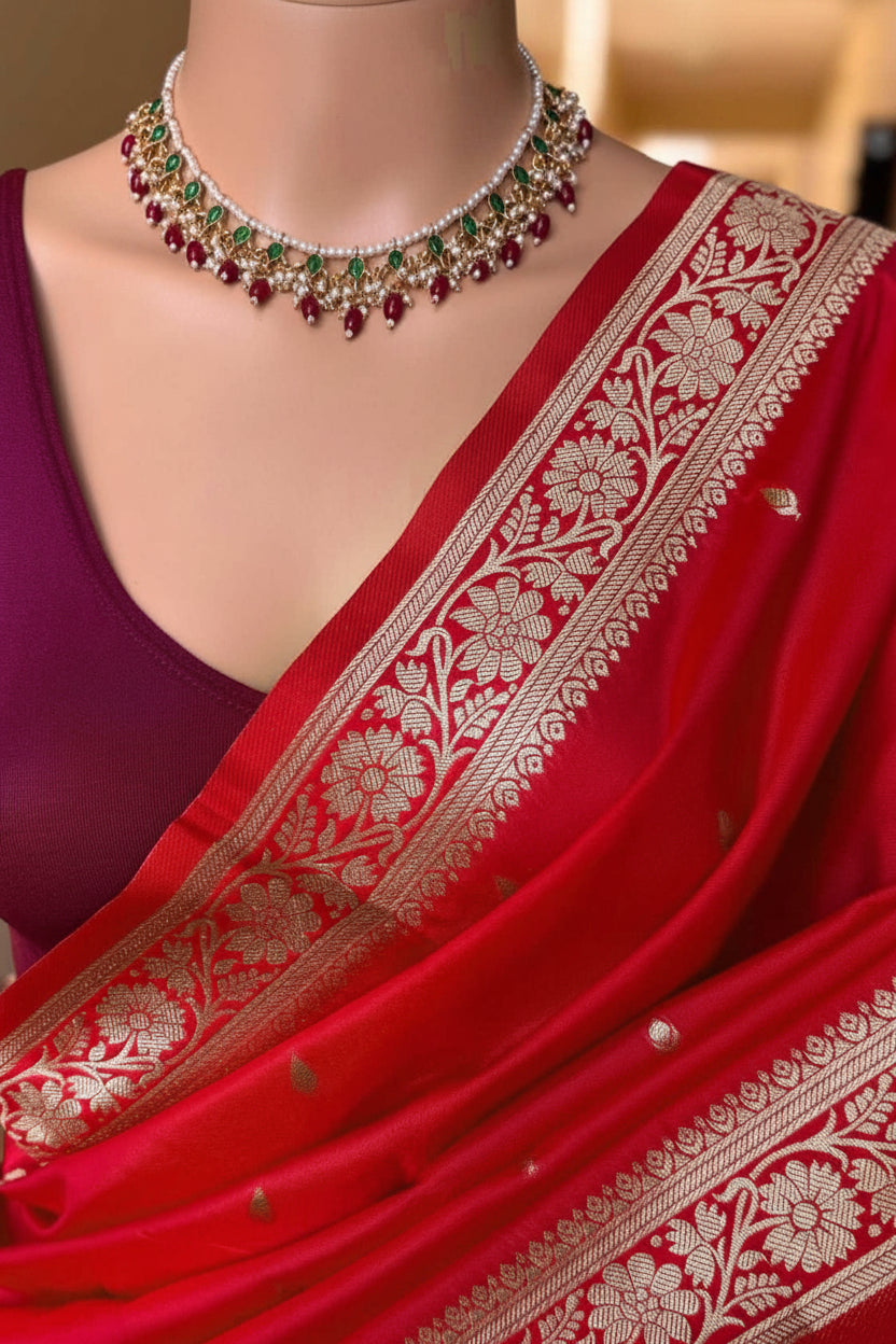 A red silk saree with intricate gold Banarasi floral zari embroidery, styled with a deep purple blouse and a matching red and green beaded necklace. The word 'Red' is printed on the left side.