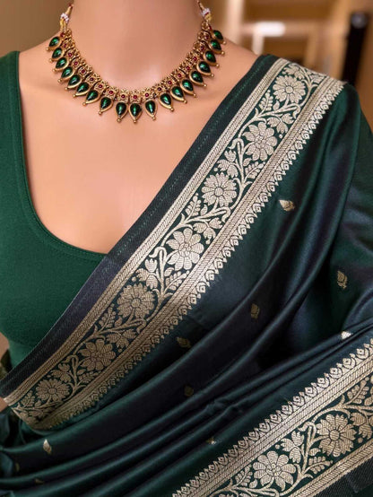 A dark green silk saree with intricate gold floral zari borders, paired with a matching dark green blouse and a gold necklace with green stones.