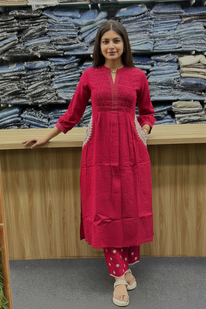 A woman takes a mirror selfie in a clothing store wearing a bright magenta-pink kurta with white embroidery on the sleeves and side pockets, paired with matching pink pants with white polka dots and beige sandals. Stacks of folded blue jeans are visible in the background.