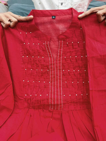 A bright red cotton kurta with white vertical embroidery and small white embroidered dots, held up by hands against a background of other garments.