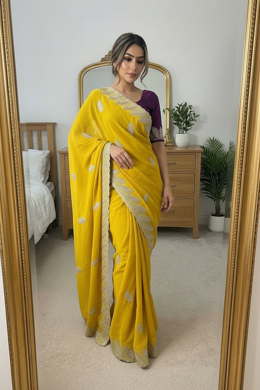 A woman takes a mirror selfie in a bedroom wearing a bright yellow sari with silver floral embroidery and a purple sleeveless blouse. The photo is taken in front of a large gold-framed mirror, with a wooden bed and dresser visible in the background.