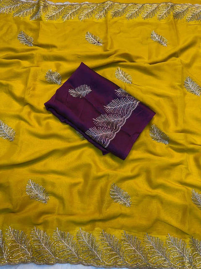 A folded deep purple fabric piece with white embroidered leaf motifs, placed on top of a bright yellow draped fabric that also features matching embroidered leaf patterns and a decorative border.
