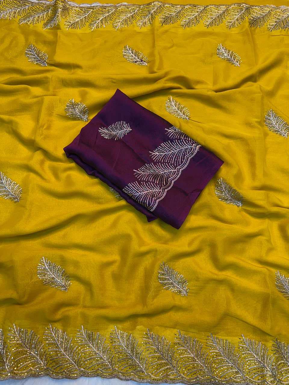 A folded deep purple fabric piece with white embroidered leaf motifs, placed on top of a bright yellow draped fabric that also features matching embroidered leaf patterns and a decorative border.