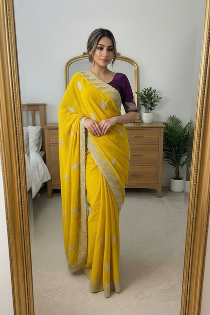 A woman takes a mirror selfie in a bedroom wearing a bright yellow sari with silver floral embroidery and a purple sleeveless blouse. The photo is taken in front of a large gold-framed mirror, with a wooden bed and dresser visible in the background.
