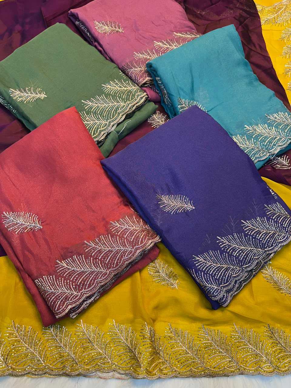 Six folded pieces of embroidered fabric in green, pink, teal, red, purple, and yellow, arranged on top of each other. Each piece features white embroidered leaf patterns and scalloped edges.