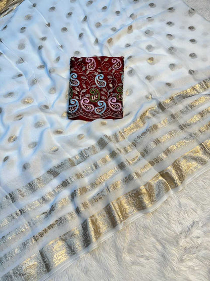 A white georgette saree with metallic gold foil dot patterns, a wide gold zari border, and a small folded red fabric patch with colorful paisley embroidery.