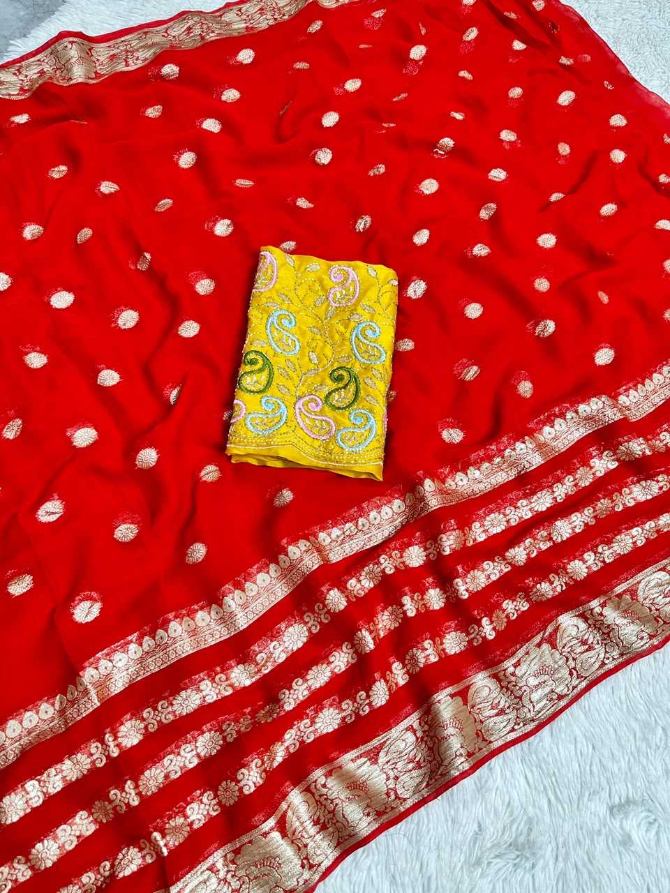 A red saree fabric with small metallic gold dots and a wide gold zari border, laid out on a white textured surface with a folded yellow patterned fabric piece placed in the center.