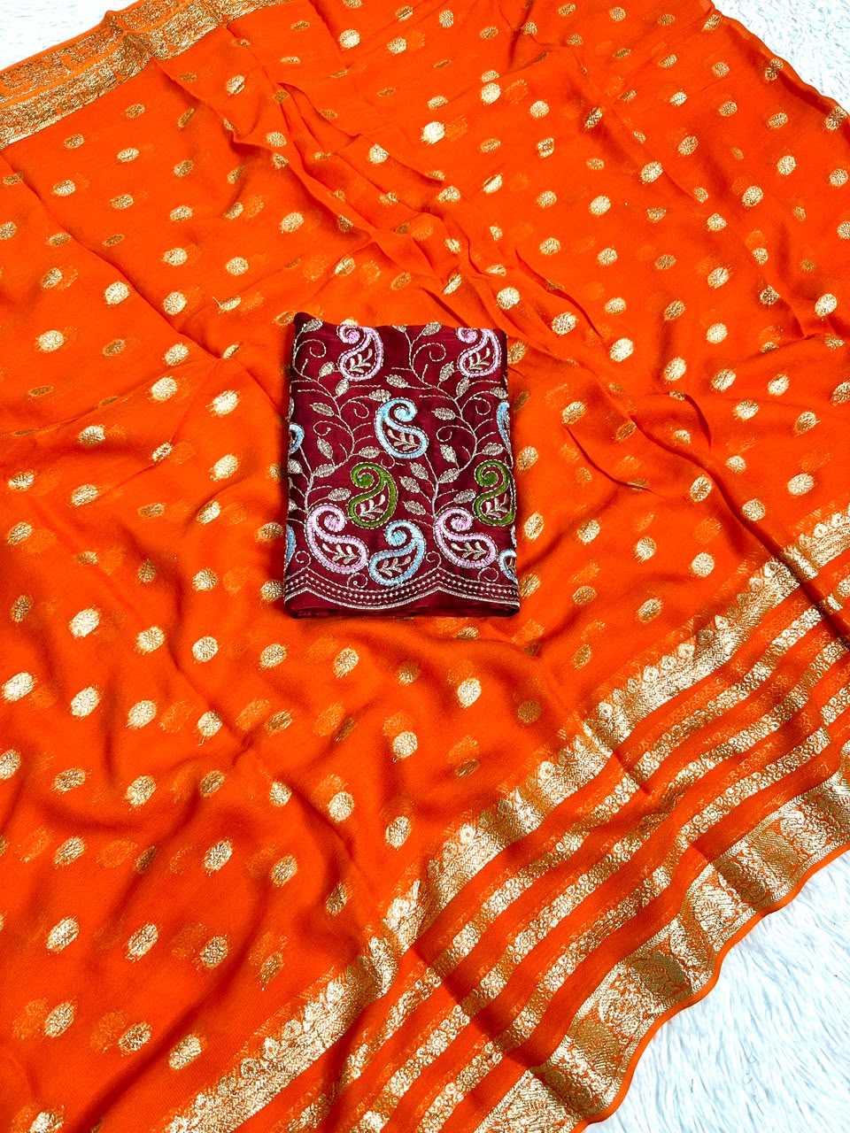 An orange saree or fabric piece with scattered gold metallic dots and a wide gold zari border, laid flat on a white textured surface. A folded matching orange fabric section with a burgundy paisley patterned center is placed in the middle.