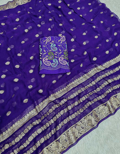 A purple georgette saree with scattered metallic gold zari motifs, a matching purple embroidered blouse piece with paisley and floral patterns, and a wide gold zari border, laid flat on a white textured surface.