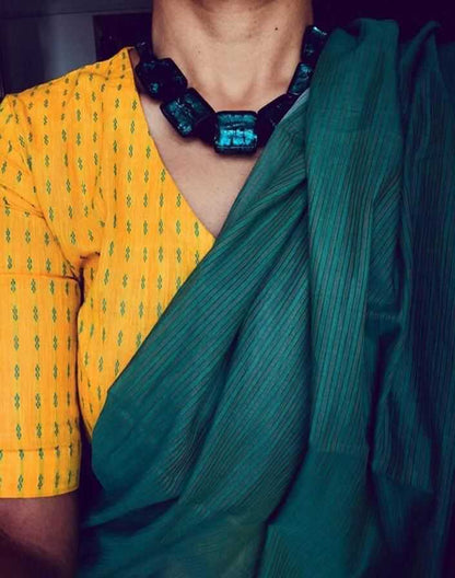 Person wearing a yellow blouse with green patterns and a green saree, accessorized with a blue necklace.
