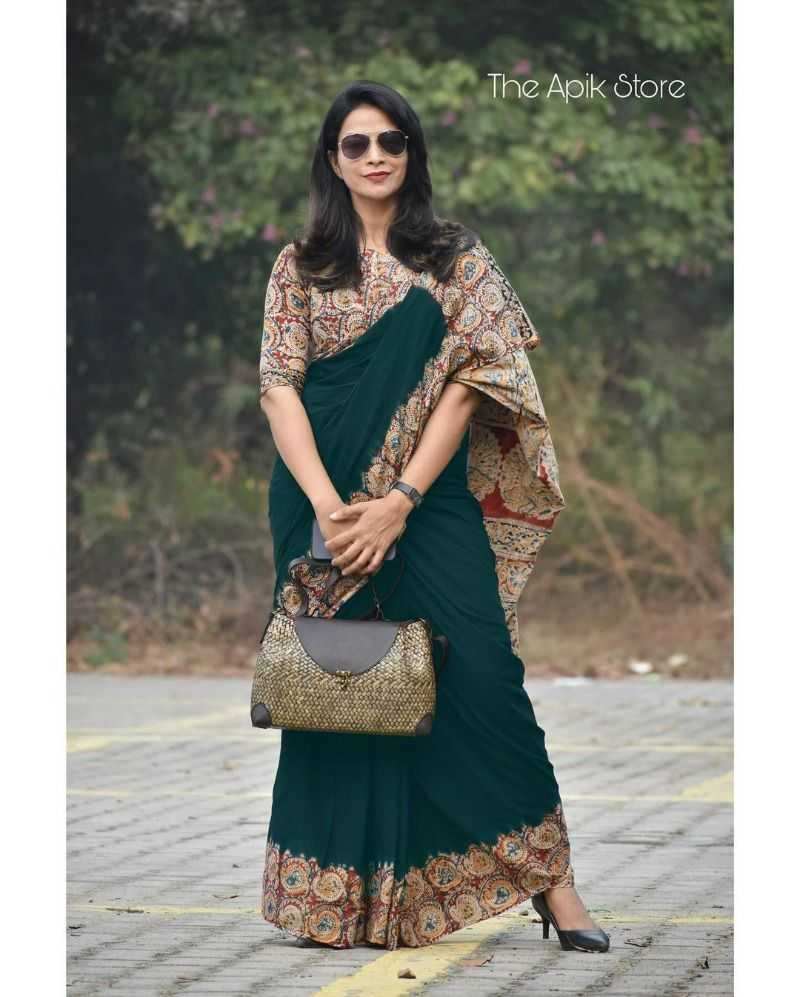 Woman in a dark green saree with a patterned blouse holding a handbag, standing outdoors.
