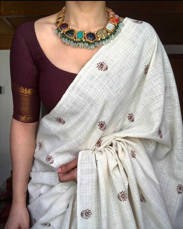 Person wearing a white saree with gold embroidery and a maroon blouse, accessorized with a colorful necklace.