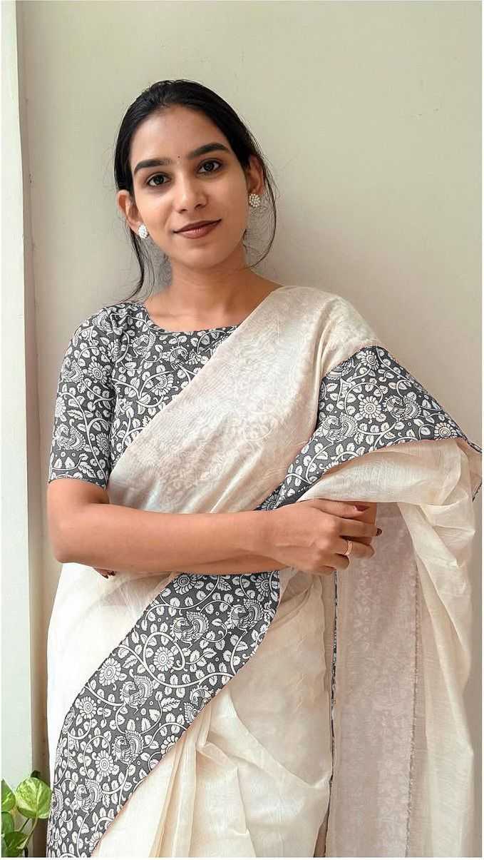 Woman wearing a white saree with a floral pattern, standing against a light green wall.