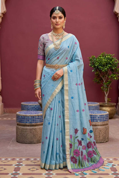 Woman in a light blue saree with floral patterns standing against a maroon wall.