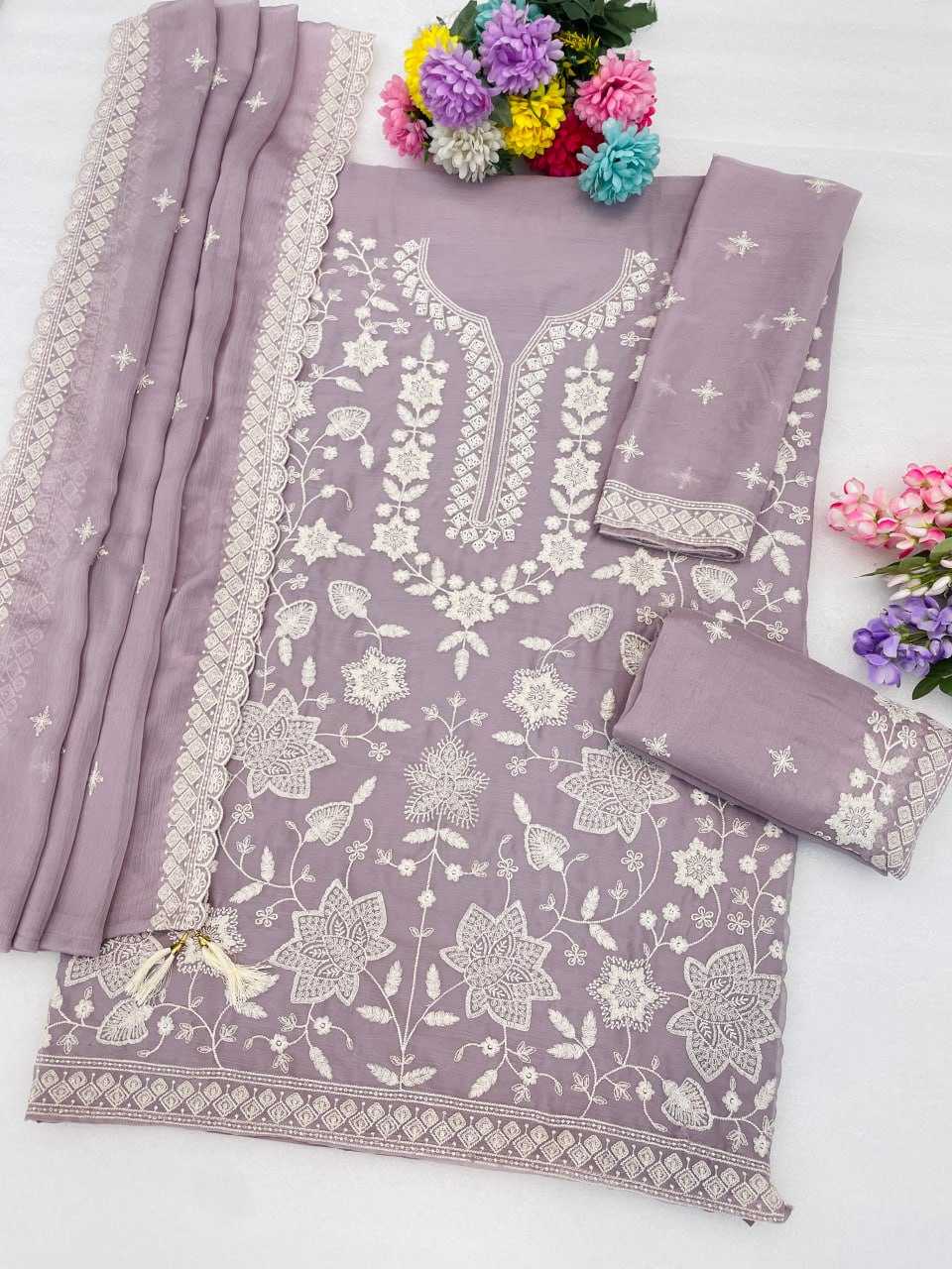 A lavender purple 3-piece salwar suit set featuring a kurta with white floral embroidery, matching trousers, and a dupatta with embroidered borders, styled with decorative flowers on a white background.