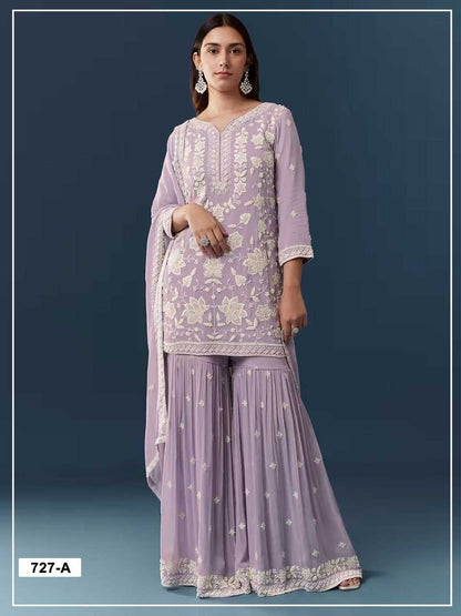 A woman modeling a lavender purple ethnic three-piece suit with beige floral embroidery, featuring a kurta, flared sharara pants, and a dupatta against a dark blue background.
