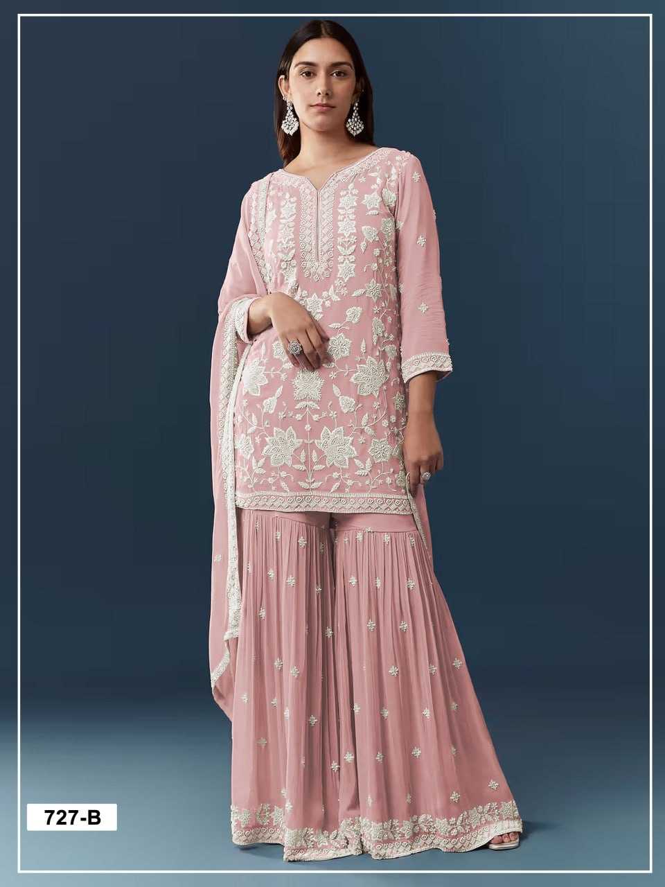 A woman modeling a pink traditional three-piece outfit with white floral embroidery, featuring a long-sleeve kurta, flared sharara pants, and a matching dupatta against a dark blue studio background.