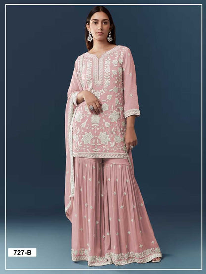 A woman modeling a pink traditional three-piece outfit with white floral embroidery, featuring a long-sleeve kurta, flared sharara pants, and a matching dupatta against a dark blue studio background.