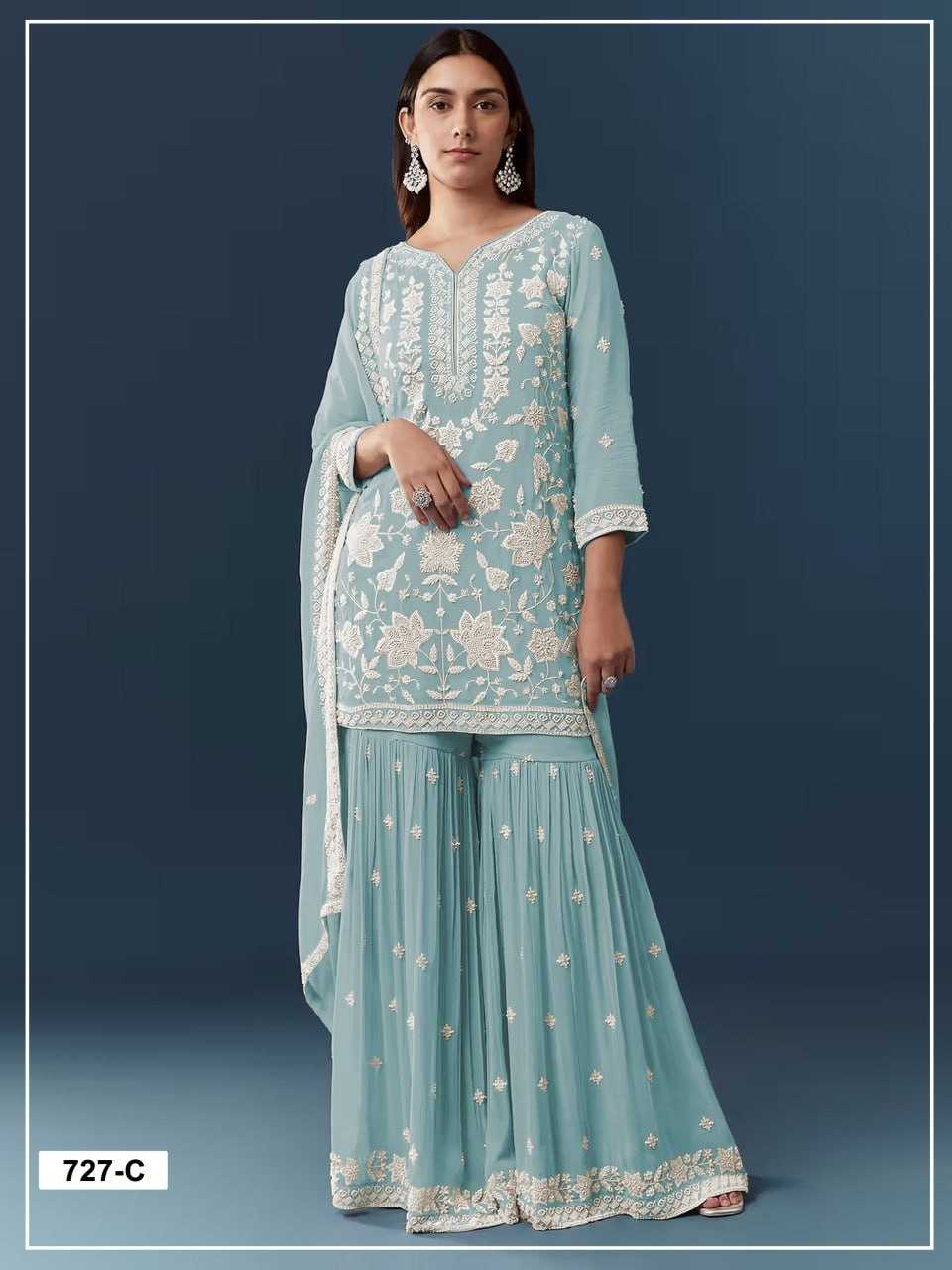 A woman modeling a light blue traditional three-piece outfit with white floral embroidery, featuring a long kurta, flared pants, and a matching dupatta against a dark blue studio background.