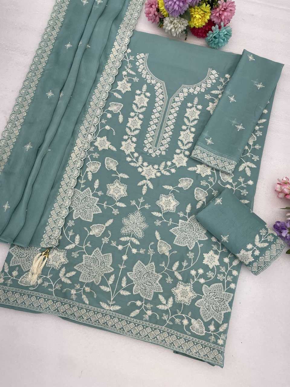 A sage green salwar suit set with intricate white floral embroidery, featuring a kurta, two matching pants, and a dupatta, styled on a light surface with decorative flowers.