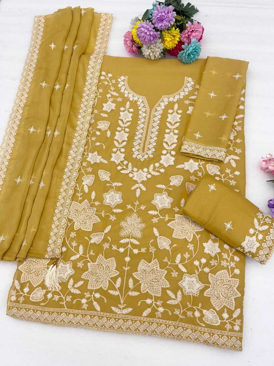 A mustard yellow ethnic suit set featuring a kurta with intricate cream floral embroidery, matching trousers, and a dupatta, styled with decorative flowers on a white background.