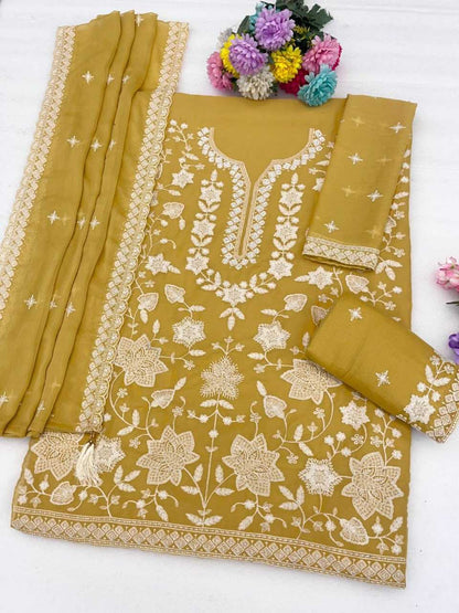A mustard yellow ethnic suit set featuring a kurta with intricate cream floral embroidery, matching trousers, and a dupatta, styled with decorative flowers on a white background.