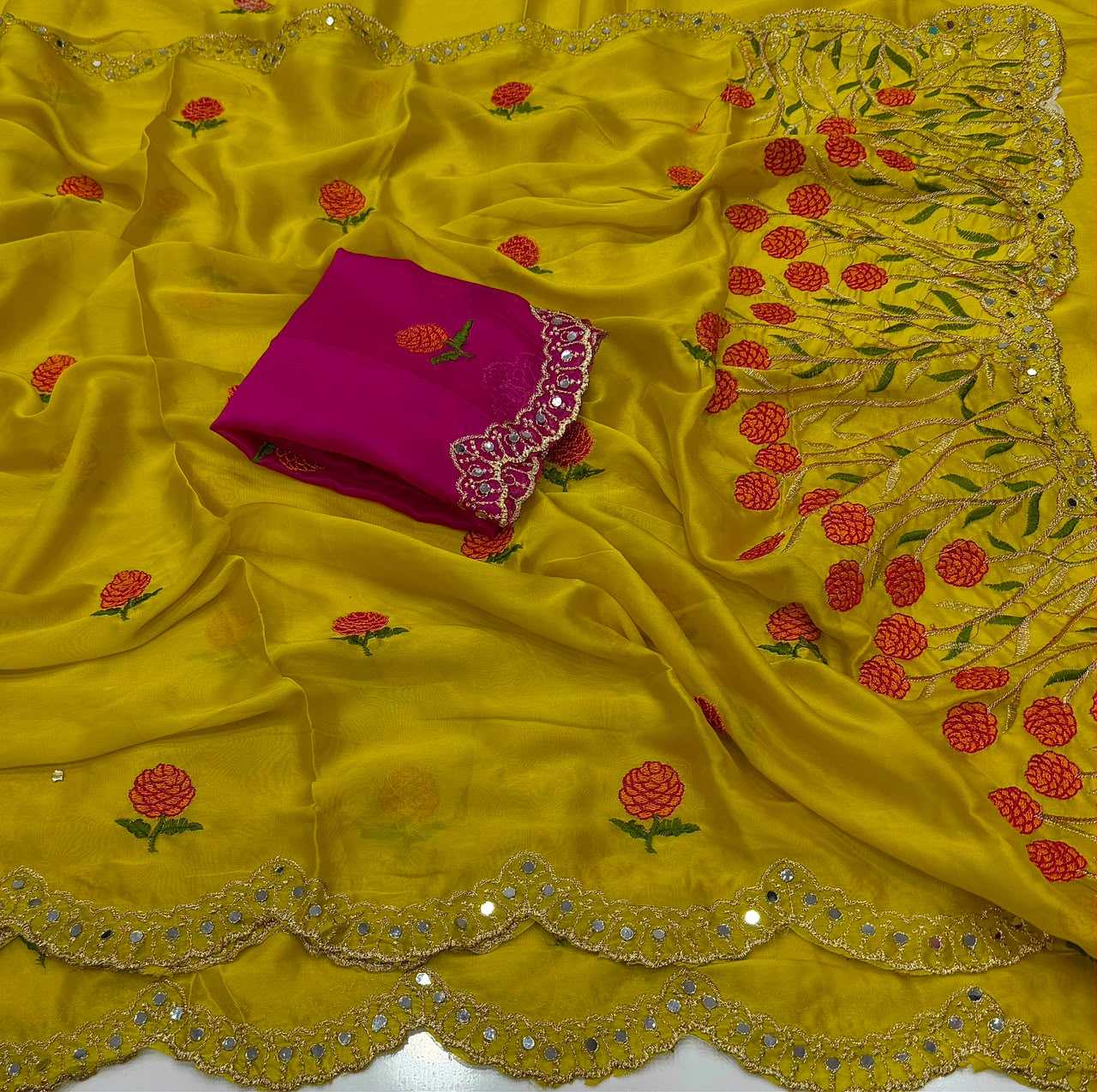 A bright yellow embroidered fabric, likely a saree or dupatta, draped to show its pattern and border details, with a folded magenta pink fabric piece featuring red and green floral embroidery and a scalloped silver mirror-work border.