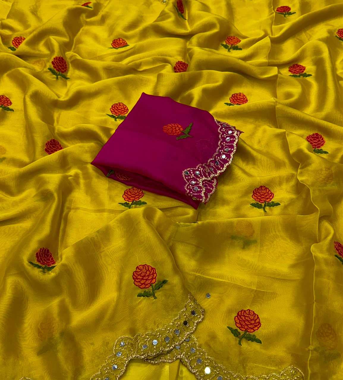 A yellow silk-like fabric with scattered red and green floral embroidery, draped to show its sheen and pattern, with a folded magenta fabric piece featuring embroidered trim placed on top.