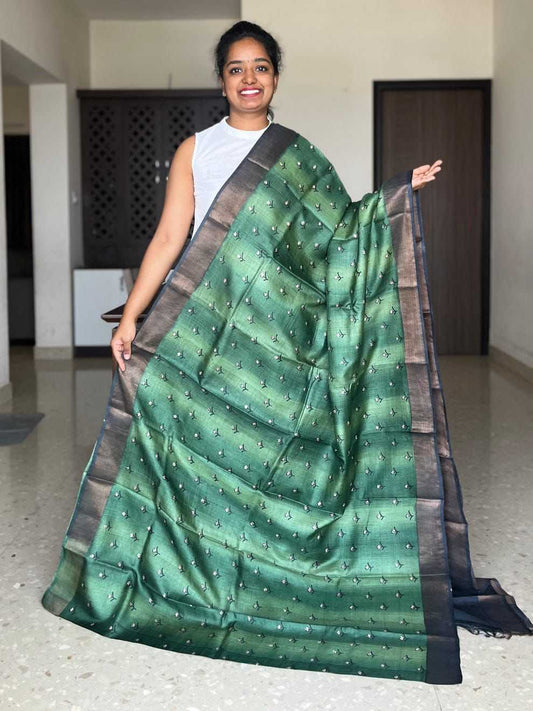 A woman modeling a green silk saree with a metallic sheen, featuring small gold motifs and a wide dark brown border, standing in an indoor hallway.