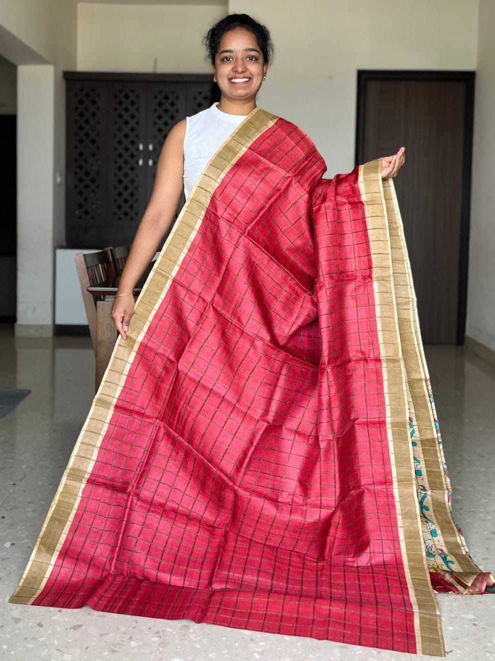 A woman wearing a red and black checkered silk saree with a gold border, holding it out to display. She is standing in an indoor room with neutral-colored walls and doors.