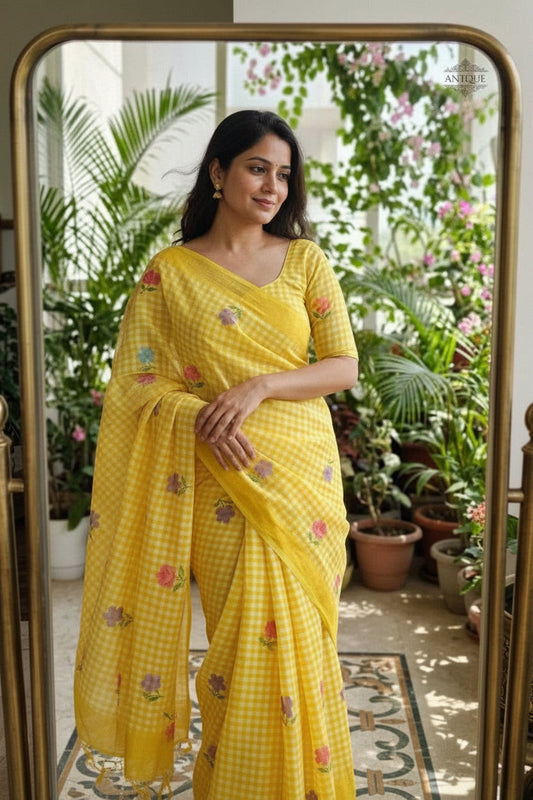 Woman taking a mirror selfie wearing a yellow saree with floral patterns in a room with plants.
