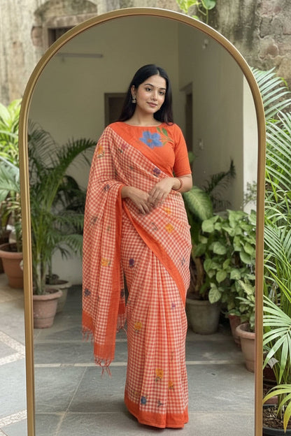 A woman takes a mirror selfie wearing a bright orange and white checkered saree with a matching embroidered blouse. The background features a large arched gold-framed mirror, potted plants, and a rustic stone wall.