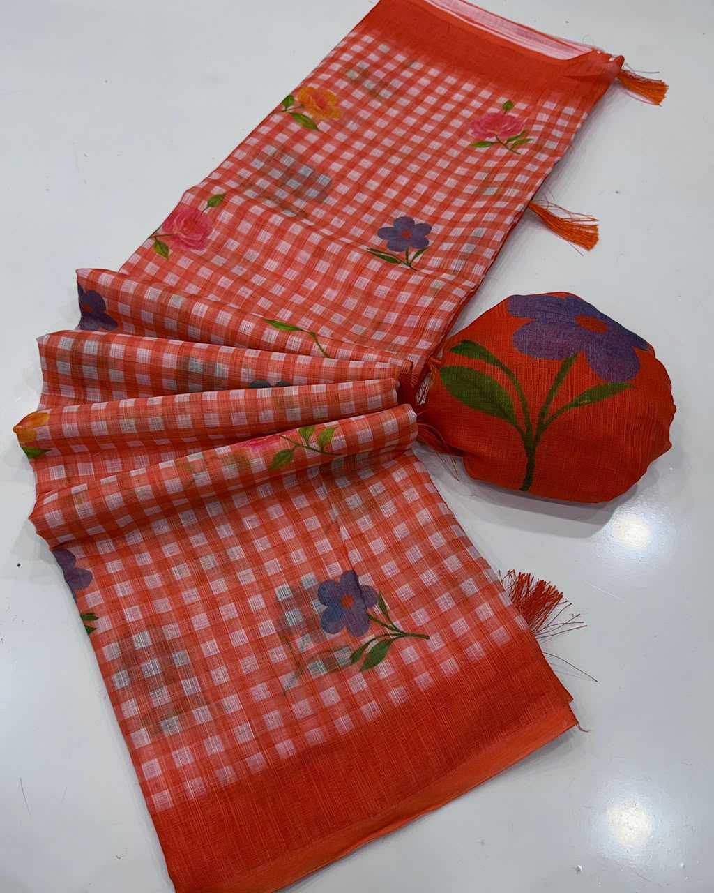 A folded orange-red checkered scarf with purple and green floral motifs, displayed next to a matching small round pouch on a white surface.