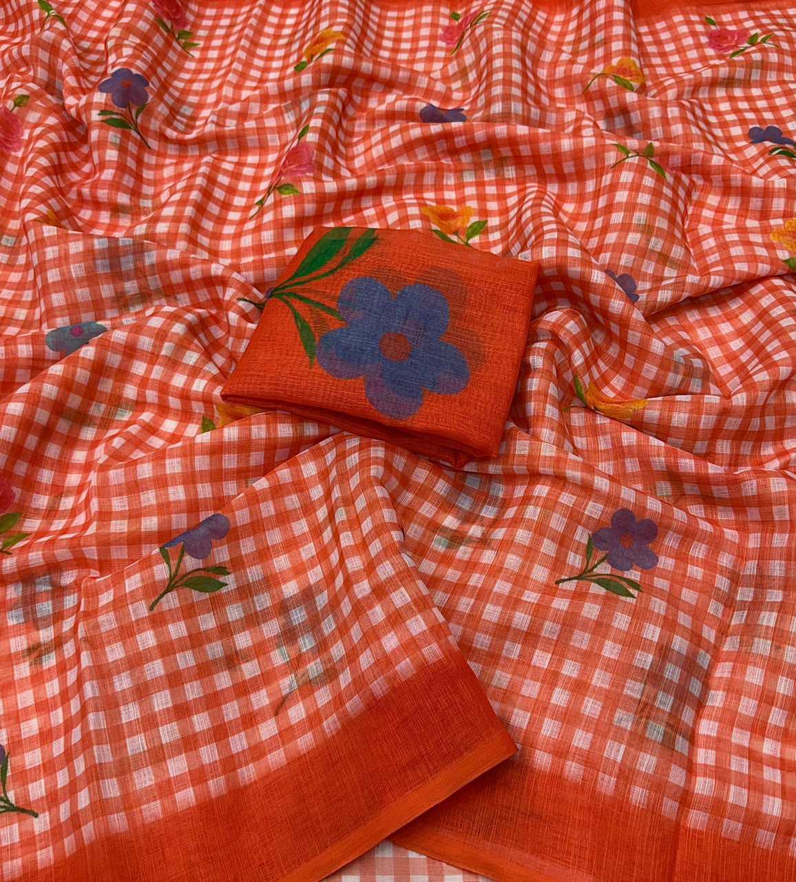 A close-up of a draped orange-red checkered fabric with scattered small floral prints in purple, green, and yellow, plus a matching solid orange-red fabric piece featuring a large blue flower and green leaves.