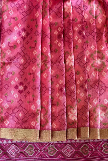 A close-up of a pink patterned fabric, likely a saree or textile, featuring vertical pleats, a gold zari border, and a matching patterned border at the bottom.