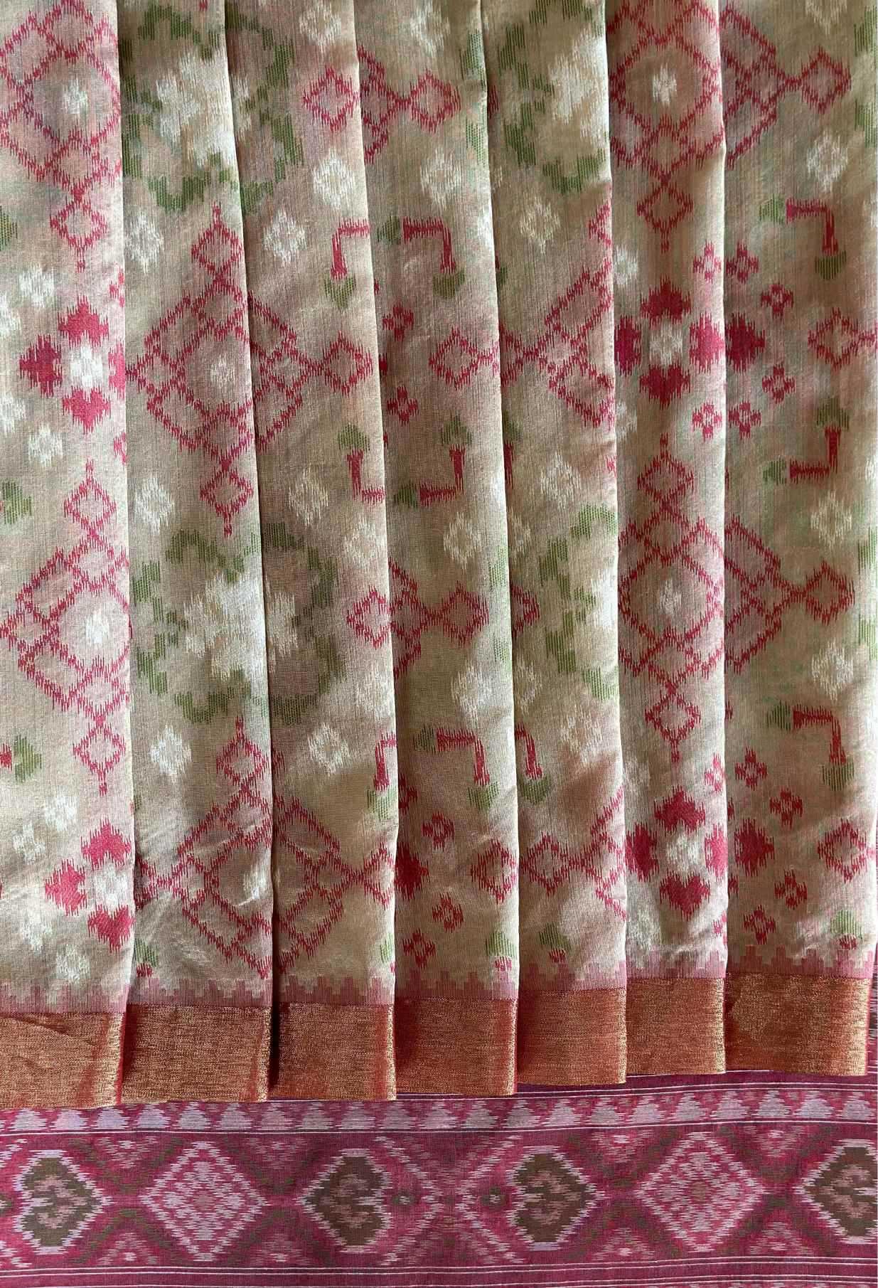 A close-up of a patterned fabric, likely a saree, featuring vertical pleats with a beige base, pink and green woven motifs, and a wide pink border at the bottom.