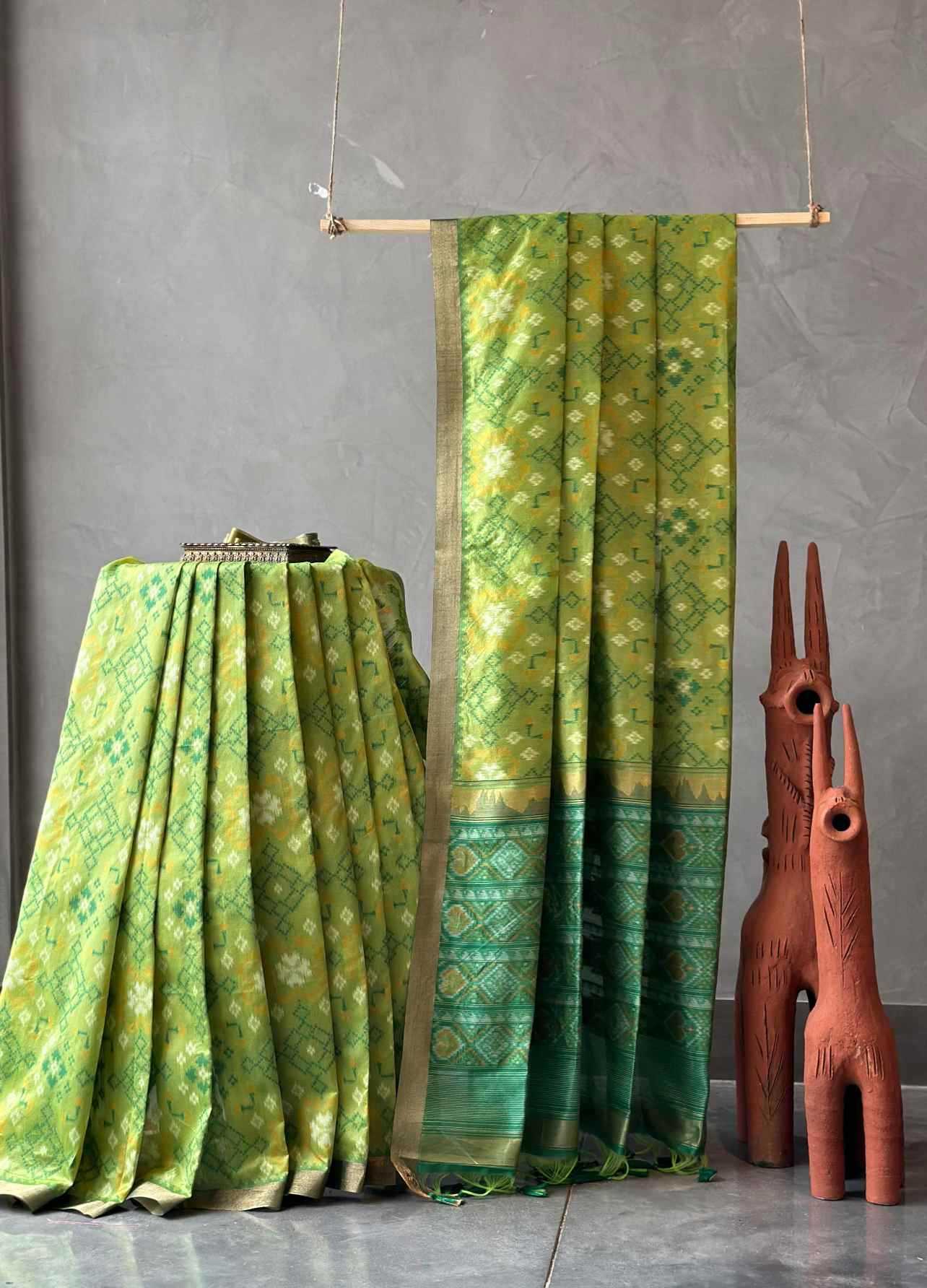 A green and yellow patterned silk saree draped elegantly, displayed alongside a matching pleated fabric piece and two terracotta decorative animal figurines against a textured gray wall.