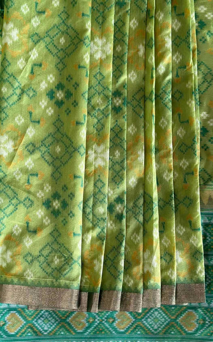 A close-up of a pleated green fabric, likely a saree, featuring a detailed geometric and floral ikat pattern in teal, yellow, and white, finished with a brown border.