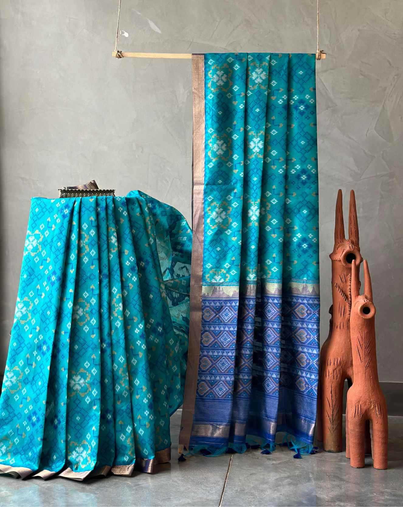 A turquoise blue patterned fabric, likely a sari or draped textile, displayed hanging from a wooden rod and draped over a stand against a textured gray wall, with two terracotta animal figurines beside it.