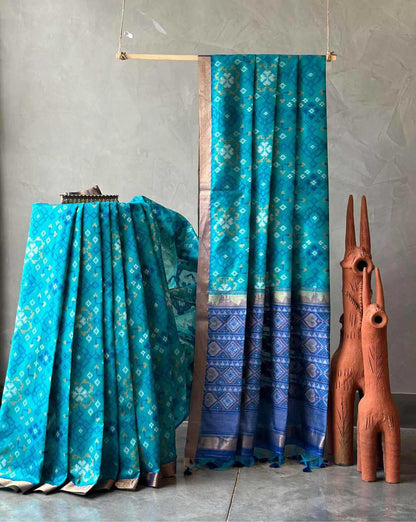 A turquoise blue patterned fabric, likely a sari or draped textile, displayed hanging from a wooden rod and draped over a stand against a textured gray wall, with two terracotta animal figurines beside it.