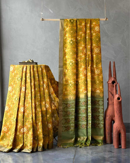 A yellow and green patterned fabric, likely a sari or large textile, draped elegantly in a studio setting. One section hangs vertically from a wooden rod, while another draped section rests on a table to the left.