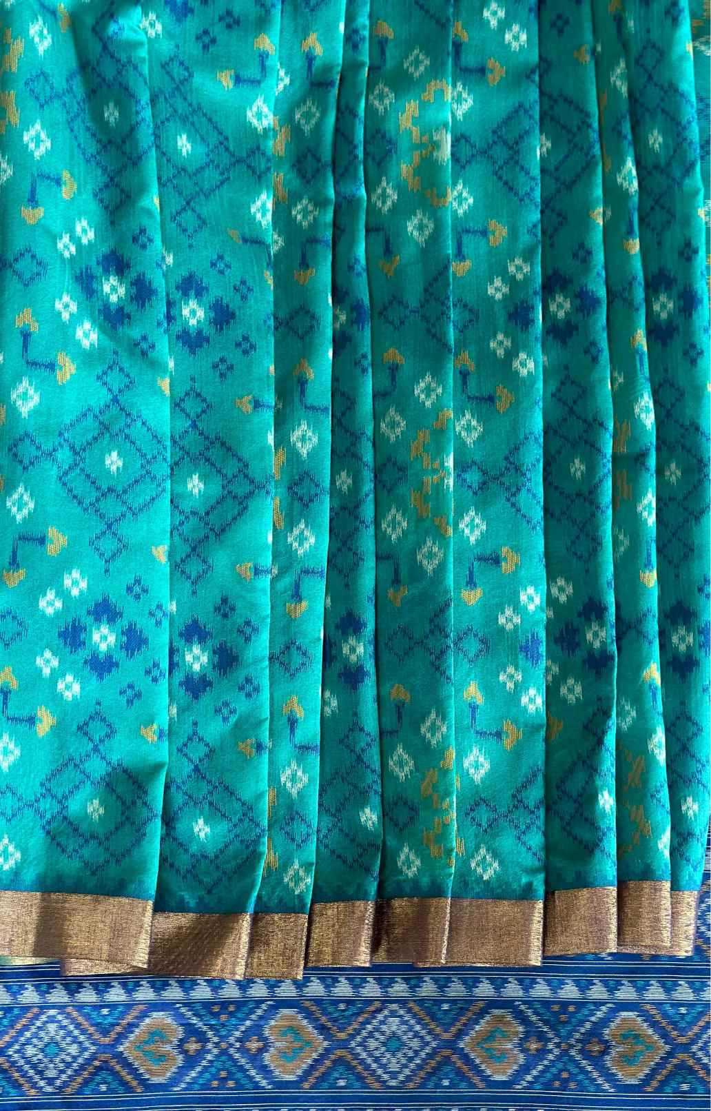 A close-up of a teal and turquoise patterned fabric, likely a sari, featuring geometric ikat-style motifs in blue, white, and gold, with a metallic gold border and a blue patterned pallu.