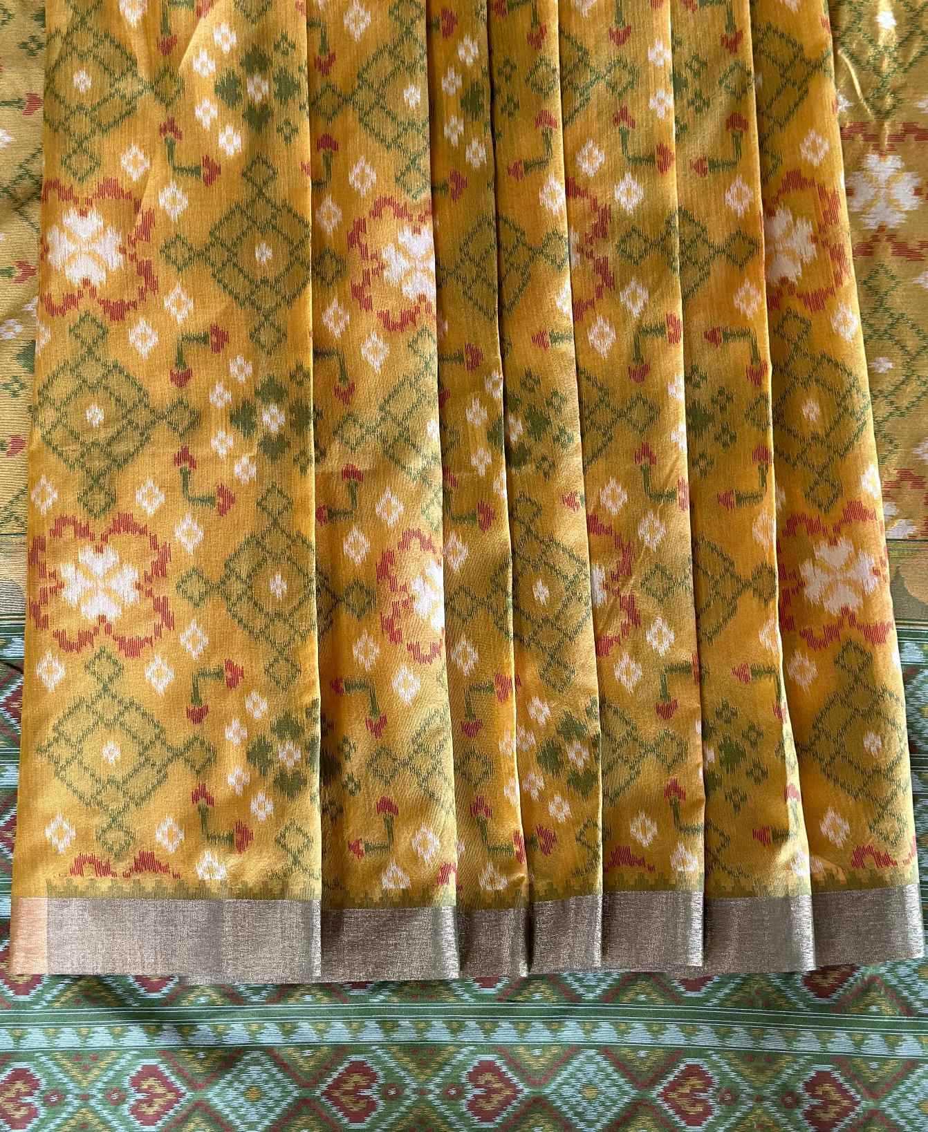 A close-up of a yellow patterned fabric, likely a saree, featuring green and red geometric and floral motifs with a metallic silver border.