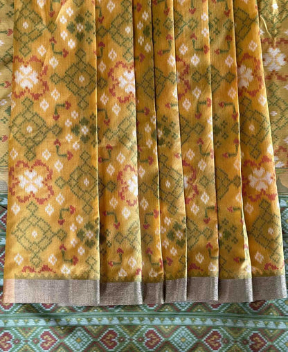 A close-up of a yellow patterned fabric, likely a saree, featuring green and red geometric and floral motifs with a metallic silver border.