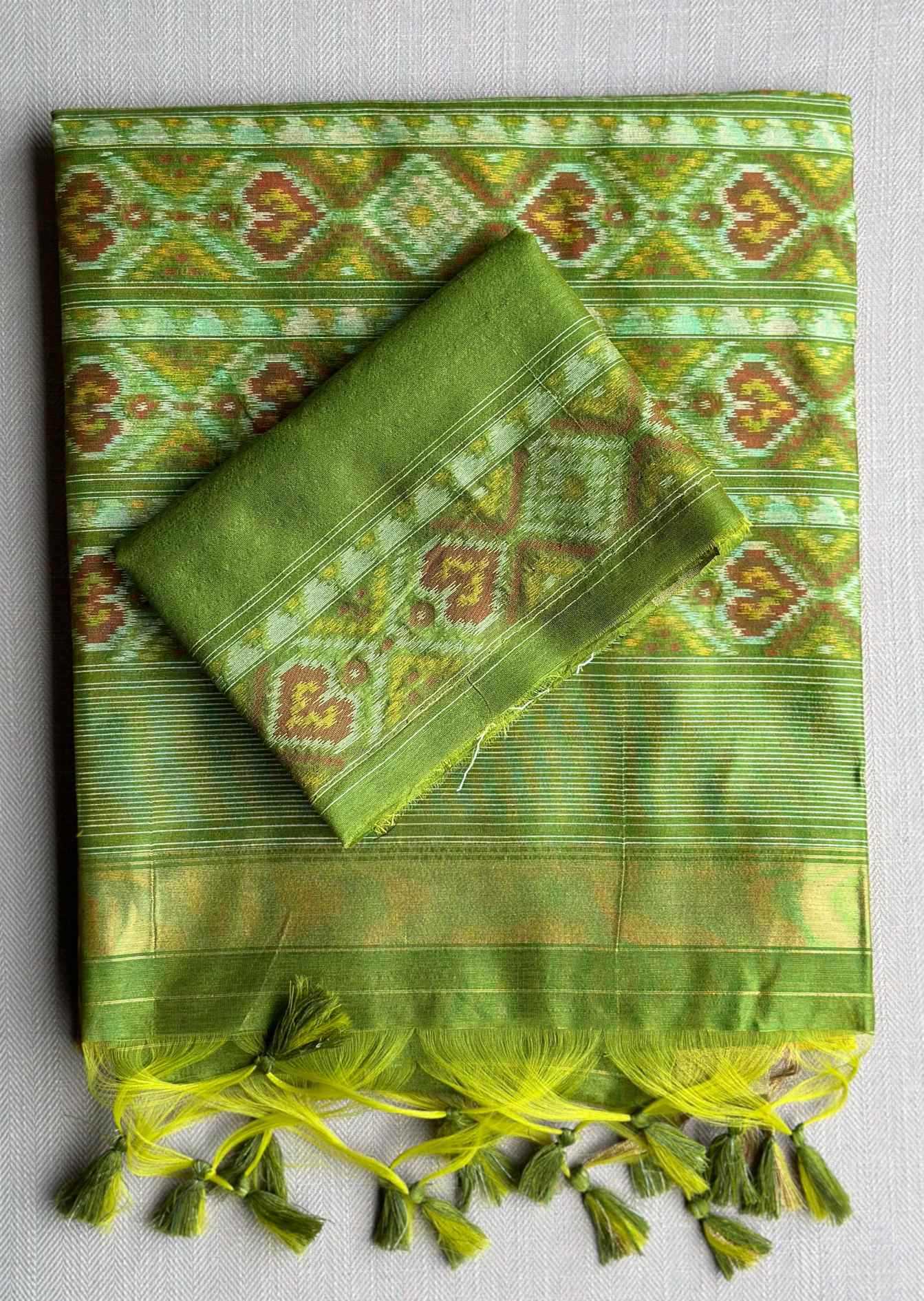 A folded green patterned fabric, likely a traditional silk saree or scarf, displayed on a neutral gray background. The fabric features intricate geometric and floral motifs in shades of green, yellow, and brown, with a matching green border and decorative tassels along the bottom edge.