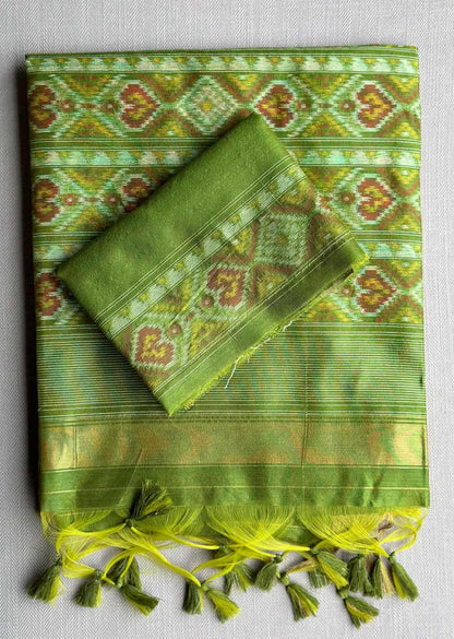 A folded green patterned fabric, likely a traditional silk saree or scarf, displayed on a neutral gray background. The fabric features intricate geometric and floral motifs in shades of green, yellow, and brown, with a matching green border and decorative tassels along the bottom edge.