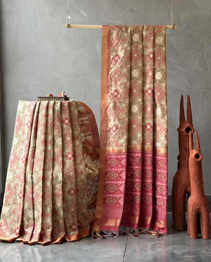 A patterned silk saree draped elegantly on a stand, with another matching drape hanging from a wooden rod against a textured gray wall. The fabric features intricate floral motifs in beige, pink, and rust red, with a wide decorative border.