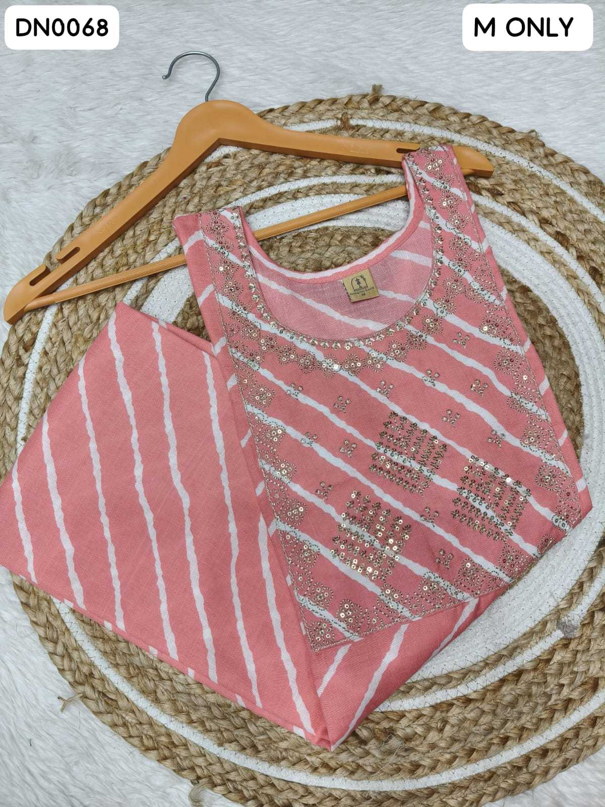A pink sleeveless kurta set with white tie-dye stripes and silver mirror work embellishments, displayed on a wooden hanger over a woven round mat.