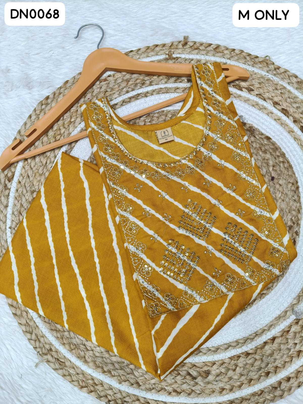 A mustard yellow sleeveless kurta top with white vertical stripes and gold sequin embroidery, displayed on a wooden hanger over a woven round mat.
