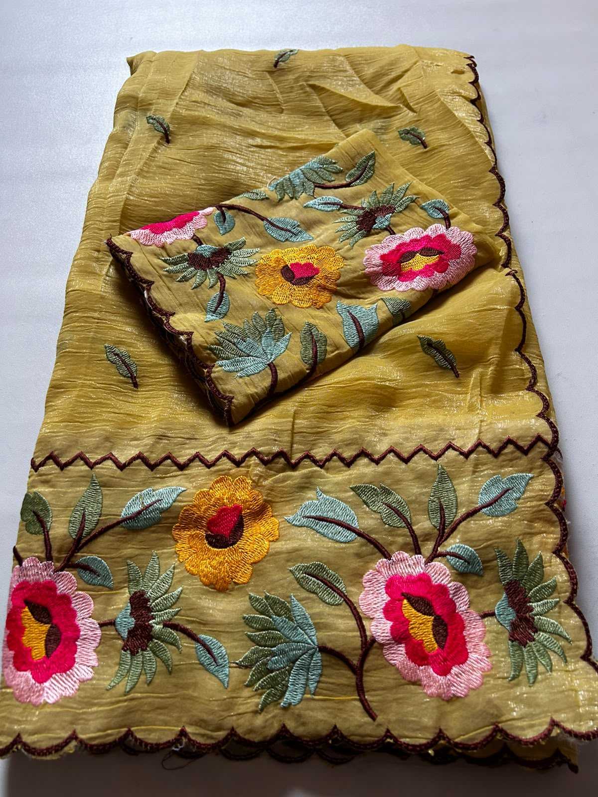 A folded yellow fabric garment or textile with embroidered floral motifs in pink, yellow, green, and brown, laid flat on a light gray background.